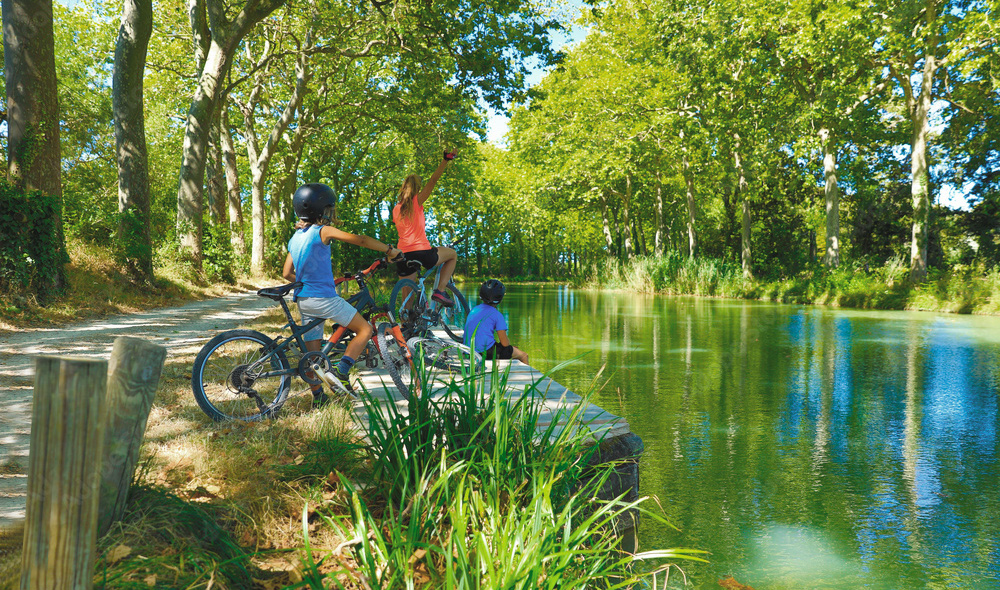 Trois enfants faisant du vélo le long d'un sentier pittoresque près d'une rivière calme, entourés d'arbres verdoyants, par une belle journée ensoleillée
