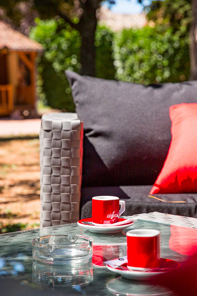 Une table en verre en plein air avec des tasses à café rouges sur des soucoupes, un cendrier, entourée d'un fauteuil avec des coussins noirs et rouges, sur fond flou d'un motel avec des bungalows et des arbres